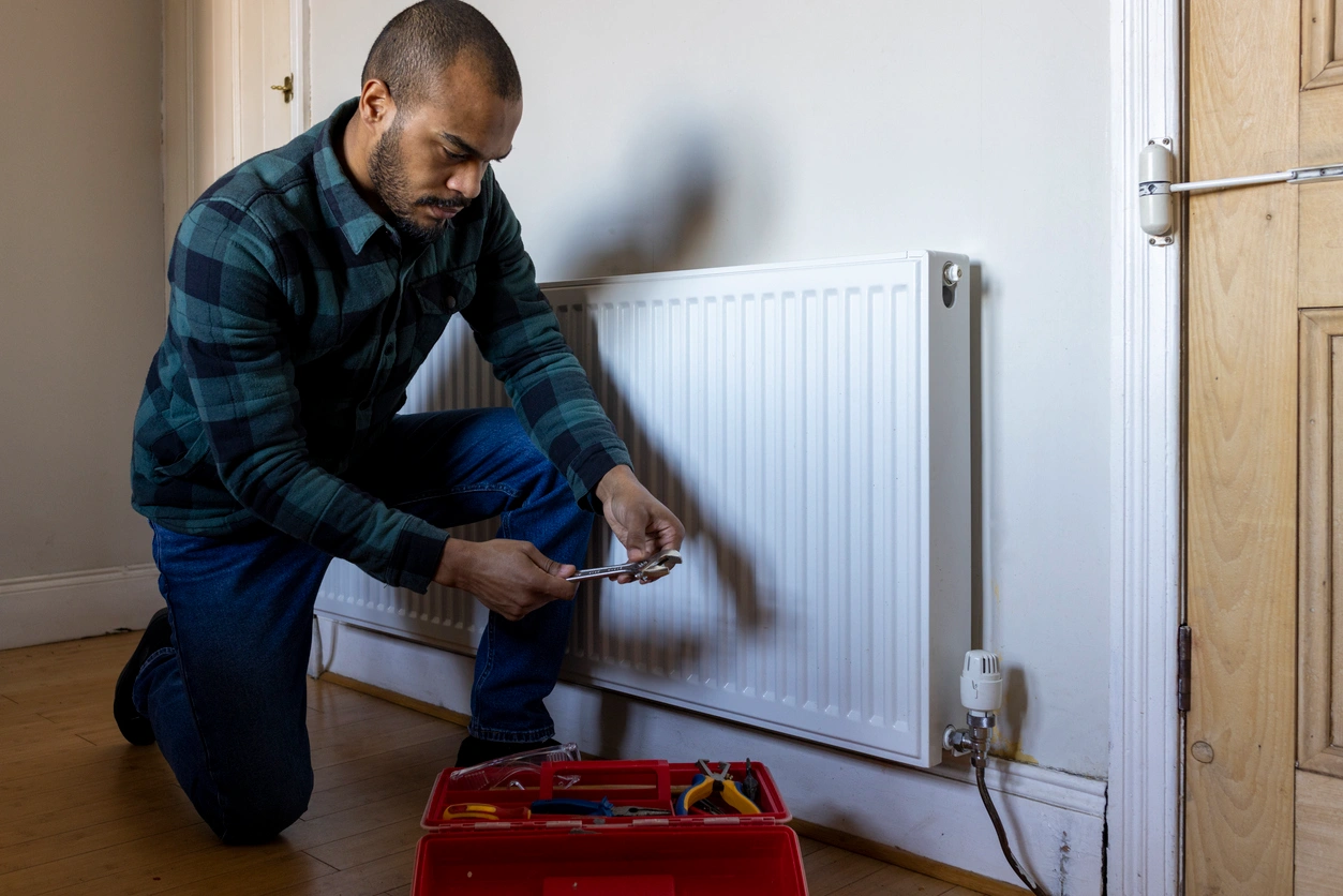 Uneven Heating Or Cooling In The House A man repairs a radiator in a room, focusing on the task with tools in hand and a determined expression.